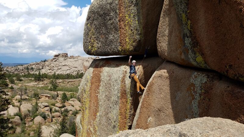 A person is rock climbing on a large rock formation in a mountainous area. The climber is wearing a helmet and harness, and is using ropes and other equipment to ascend the rock face. The rock formation is composed of large, rounded boulders, and the surrounding landscape is characterized by trees, grass, and other vegetation. The sky is partly cloudy.